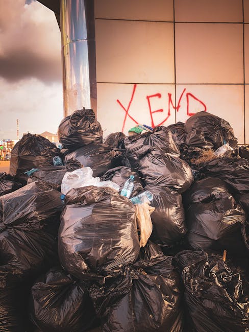 A large pile of black rubbish bags, some with visible creases and tied at the top, is stacked against a partly reflective, metallic surface on an urban street scene. The bags are filled with waste and