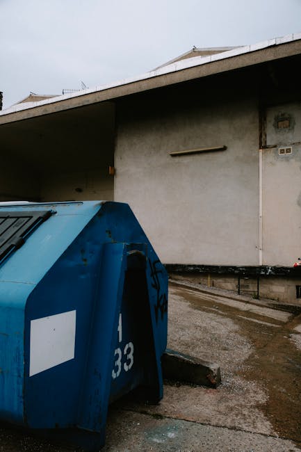 A large, blue, industrial-style waste container with a curved, ribbed metal surface and a white rectangular label, positioned on a paved outdoor area adjacent to a plain, beige concrete building wall.